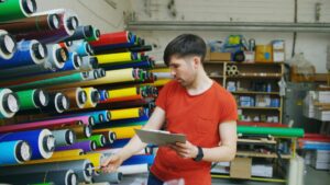 Technician selecting rolls of colorful marine vinyl wrap material in a workshop while checking inventory on a clipboard