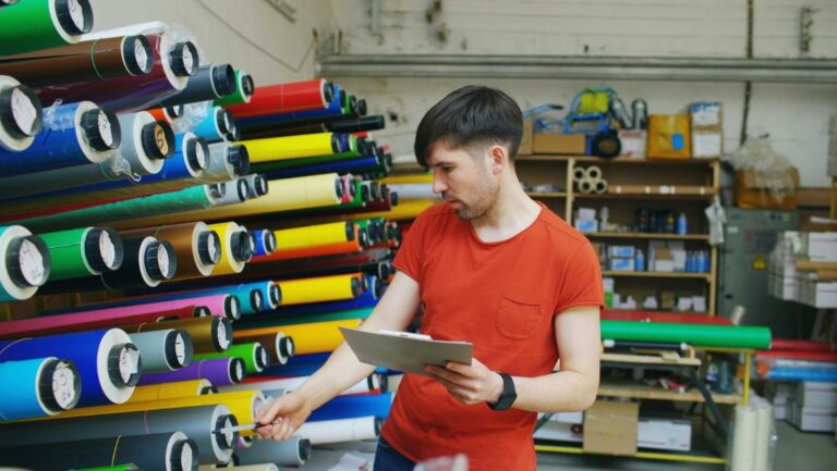 Technician selecting rolls of colorful marine vinyl wrap material in a workshop while checking inventory on a clipboard