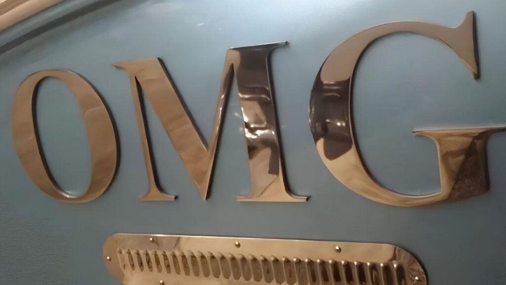 Polished stainless steel “OMG” boat lettering mounted on a light blue hull above a vent.